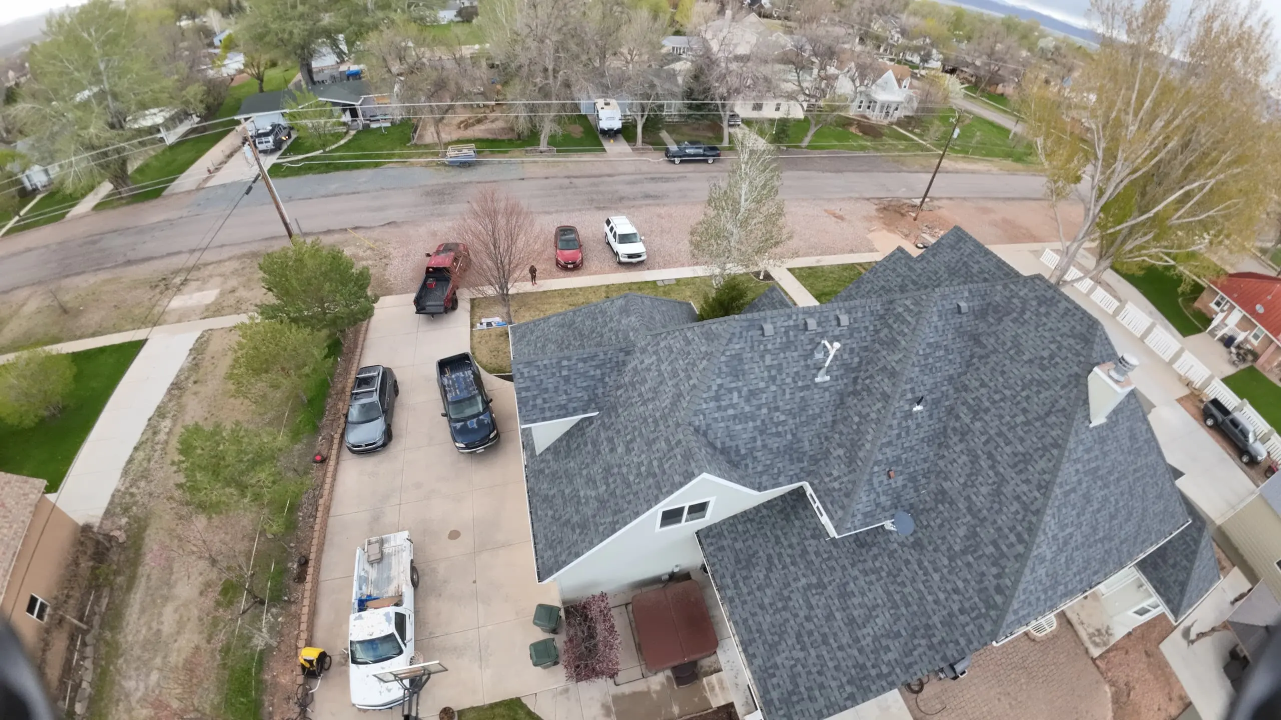 Aerial view of a residential neighborhood showing several houses with large yards. The focus is on a gray-roofed house with multiple cars parked in the driveway and along the street. Trees and green lawns are visible, and the area appears peaceful and well-maintained.