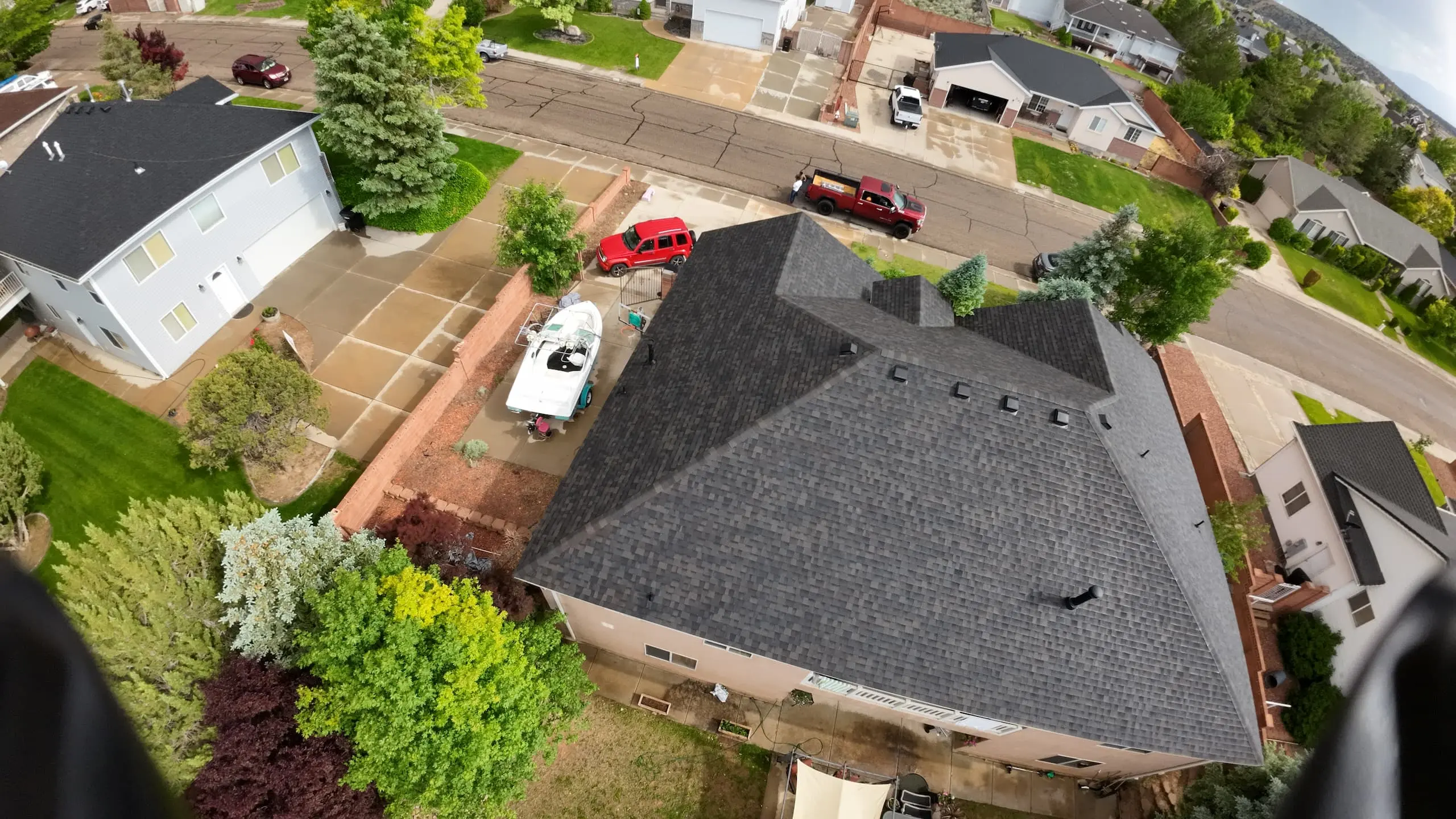 Aerial view of a residential neighborhood showing a house with a dark shingled roof and a boat parked in the driveway. The house has a large, well-maintained yard with various trees and shrubs. Surrounding houses also feature spacious driveways and green lawns. Several cars are parked on the street and in the driveways. The area appears peaceful and suburban, with neatly arranged homes and greenery.