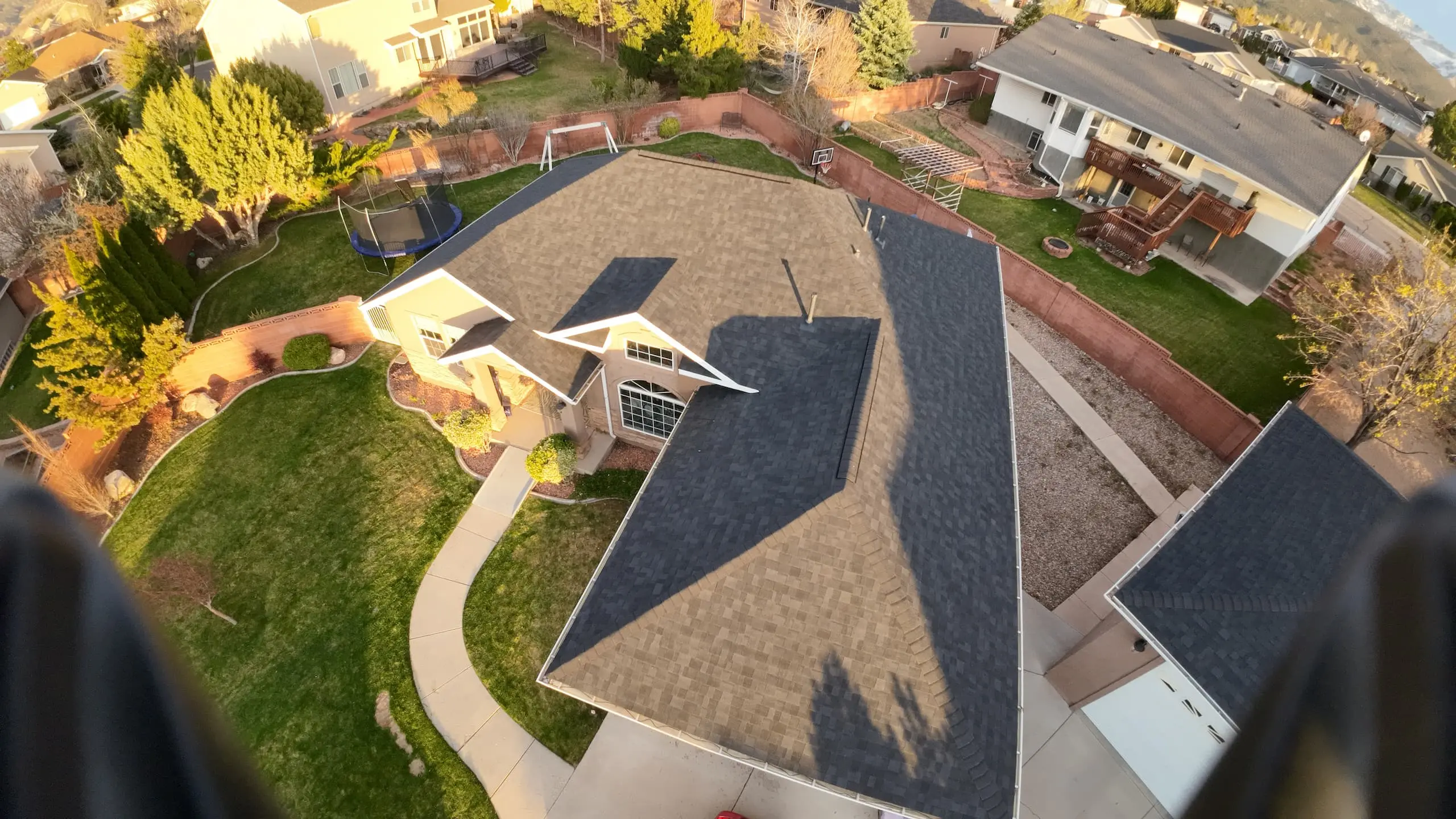 Aerial view of a residential neighborhood focused on a house with a dark shingled roof and a large backyard. The yard includes a well-manicured lawn, a trampoline, a basketball hoop, and several trees and shrubs. The surrounding area shows other houses with spacious yards and greenery, set against a backdrop of mountains in the distance.