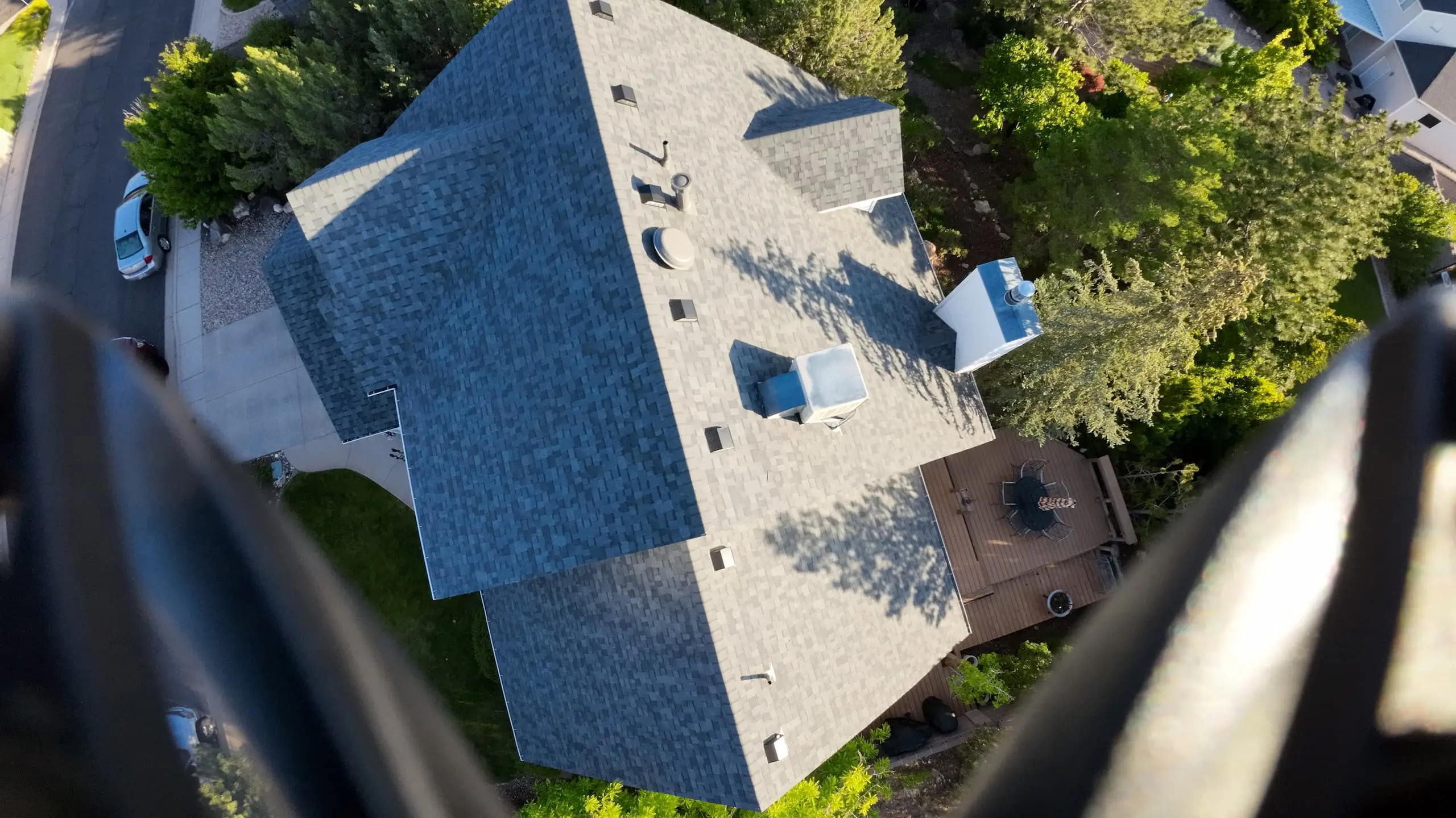 Aerial view of a house with a dark shingled roof, surrounded by lush green trees and bushes. The driveway has several cars parked, and the backyard features a wooden deck with outdoor furniture. The neighborhood has a variety of houses with well-maintained lawns and greenery.