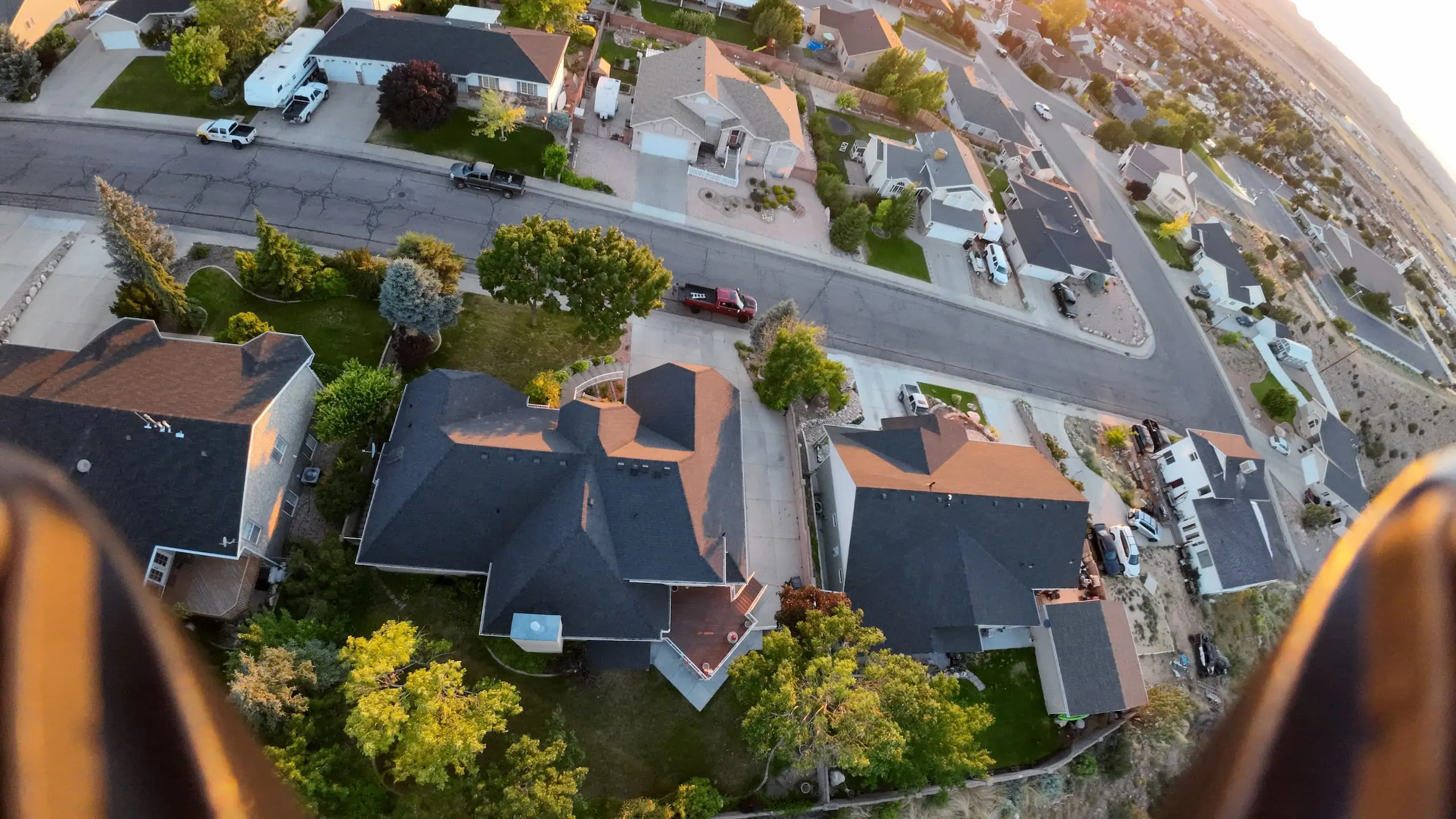 Aerial view of a neighborhood showing a house with a large, dark shingled roof and a well-kept yard. Several cars are parked in the driveway and on the street. The area is green with many trees and well-maintained lawns. Other houses with similar characteristics are visible in the vicinity.