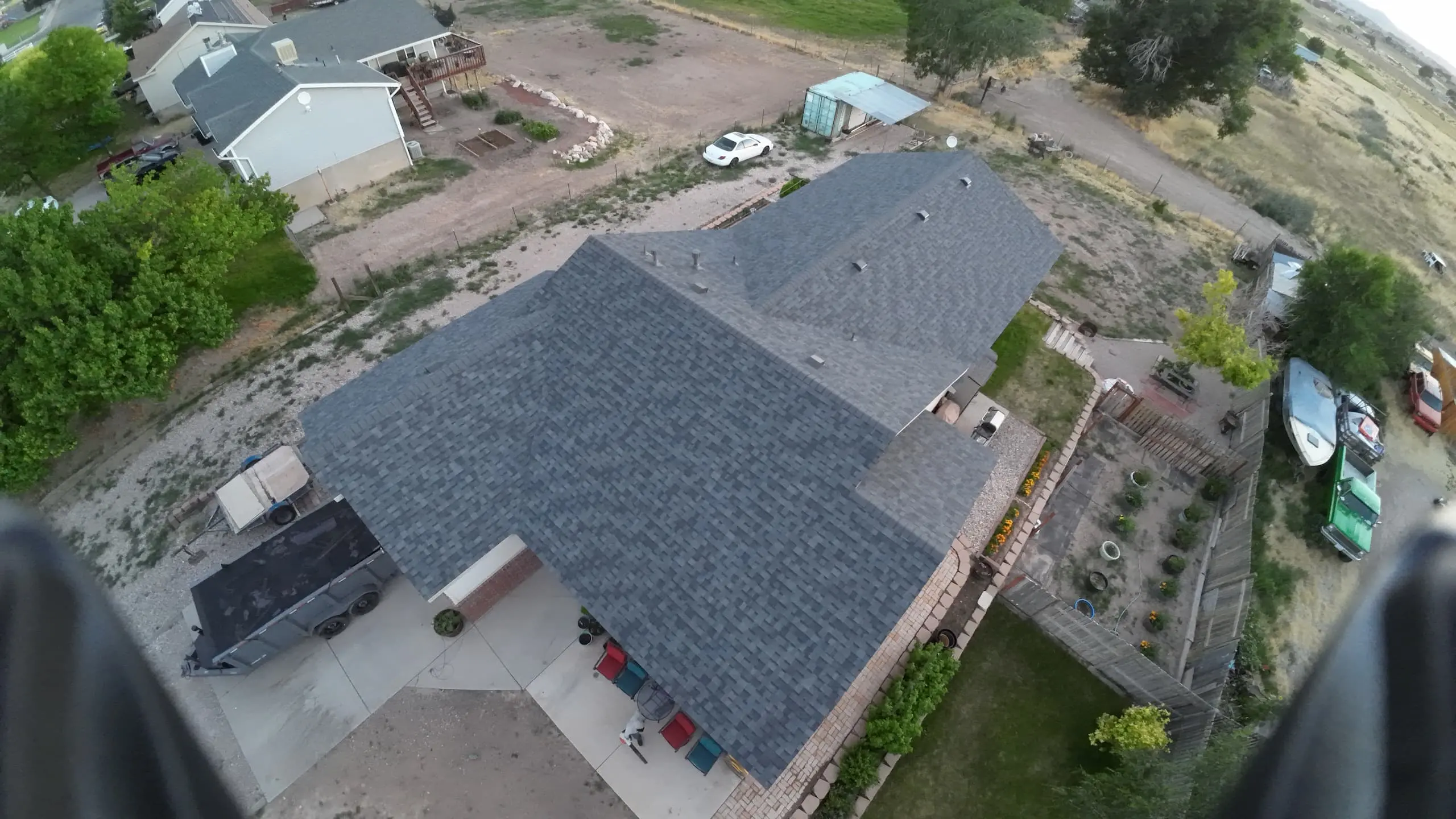 Aerial view of a suburban neighborhood with a focus on a house with a light gray shingled roof. The driveway has a car and a trailer parked, and the backyard is well-maintained with various plants and trees. Surrounding houses have similar features, with green lawns and trees.