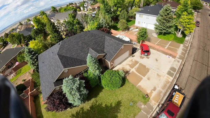 Aerial view of a house with a dark shingled roof and a manicured lawn. The driveway has a red car parked, and the yard is surrounded by trees and shrubs. The neighborhood features similar houses with green lawns and a mix of trees and plants.